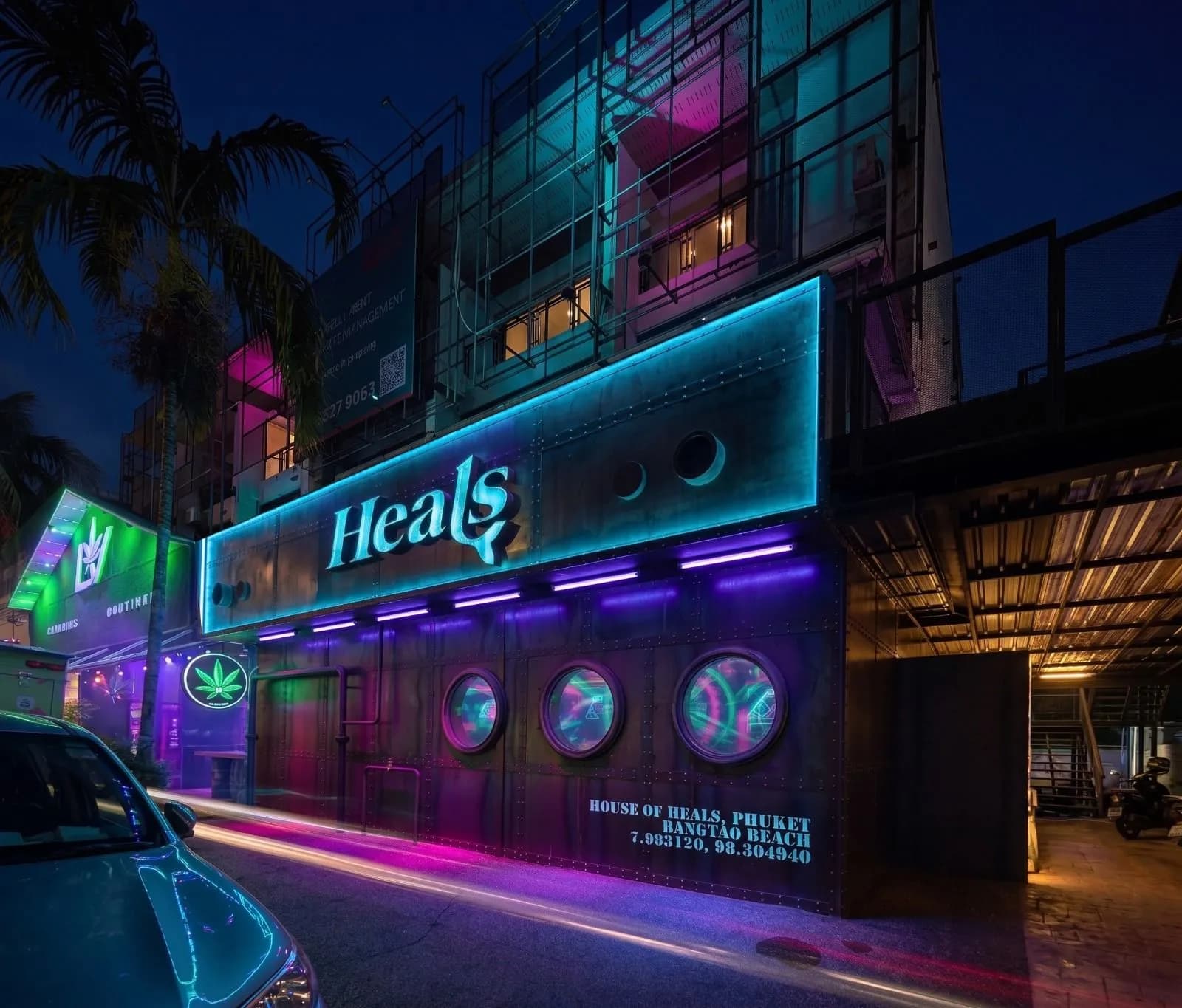 The Heals Phuket façade at night: riveted steel, the wordmark lit teal and portholes glowing magenta